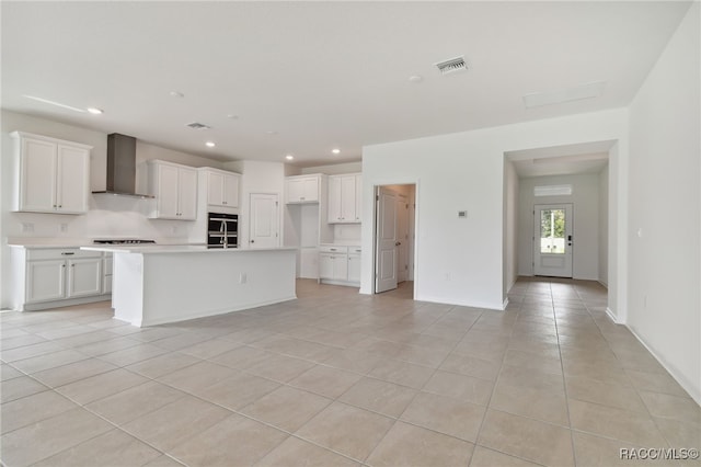 kitchen with white cabinetry, wall chimney range hood, stovetop, an island with sink, and light tile patterned floors