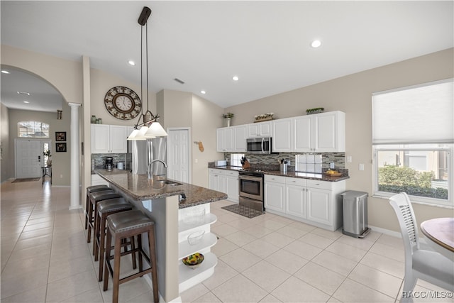 kitchen featuring ornate columns, stainless steel appliances, dark stone countertops, decorative light fixtures, and a kitchen island with sink