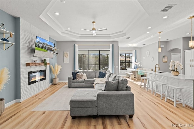 living room featuring a raised ceiling, ceiling fan, a large fireplace, and light wood-type flooring