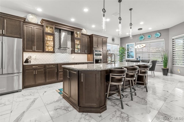 kitchen with light stone countertops, appliances with stainless steel finishes, wall chimney exhaust hood, a kitchen island with sink, and hanging light fixtures