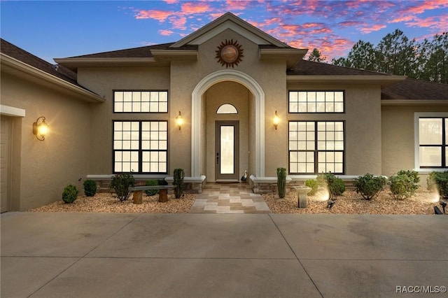 exterior entry at dusk featuring an attached garage, a shingled roof, and stucco siding