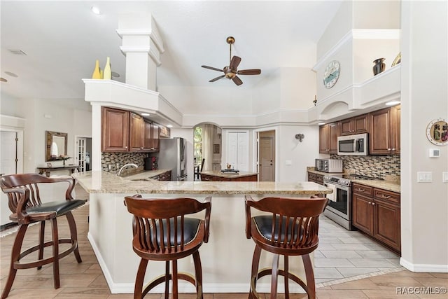 kitchen with kitchen peninsula, decorative backsplash, a towering ceiling, appliances with stainless steel finishes, and a kitchen breakfast bar