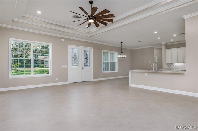 unfurnished living room with ceiling fan with notable chandelier, a sink, a tray ceiling, crown molding, and baseboards