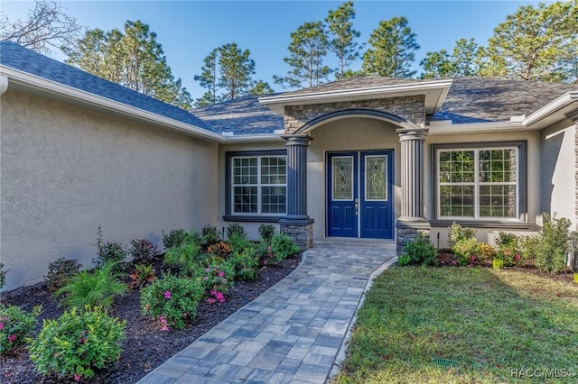 doorway to property featuring stone siding, stucco siding, a lawn, and a shingled roof