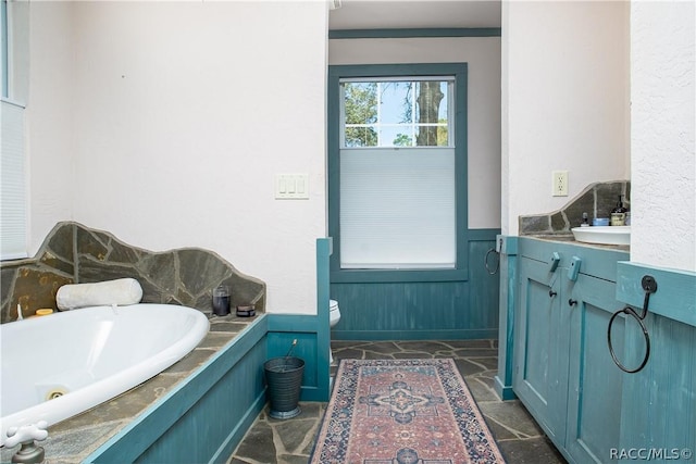 bathroom featuring a wainscoted wall, a garden tub, toilet, stone finish flooring, and vanity