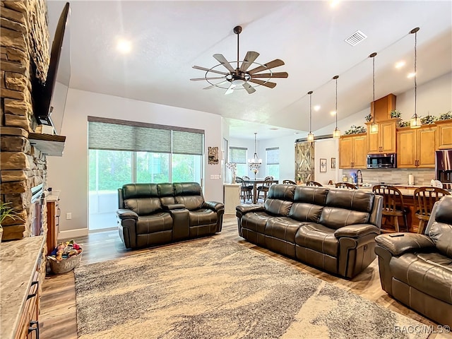 living room featuring ceiling fan, sink, high vaulted ceiling, light hardwood / wood-style floors, and a stone fireplace