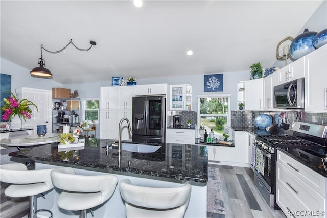 kitchen featuring light wood-type flooring, stainless steel appliances, decorative light fixtures, a center island with sink, and white cabinets