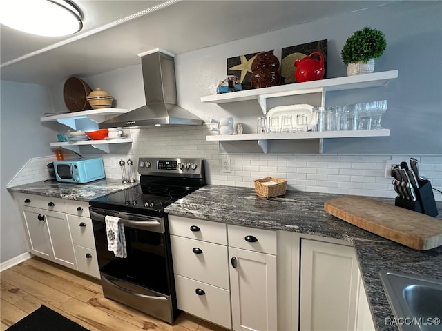 kitchen featuring wall chimney range hood, stainless steel range with electric stovetop, backsplash, light hardwood / wood-style floors, and white cabinets