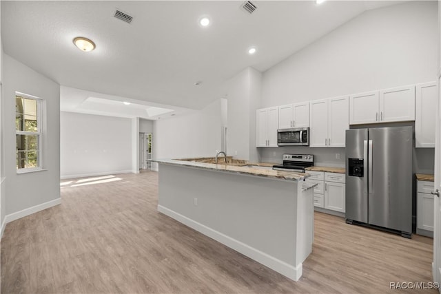 kitchen with white cabinets, appliances with stainless steel finishes, a kitchen island with sink, light wood-type flooring, and light stone counters