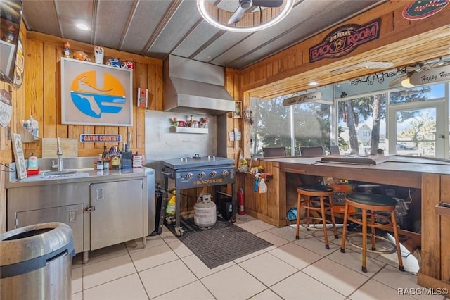 kitchen featuring wall chimney range hood, sink, wooden walls, ceiling fan, and light tile patterned flooring