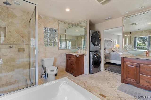 bathroom with stacked washer / dryer, toilet, vanity, a shower with shower door, and tile walls