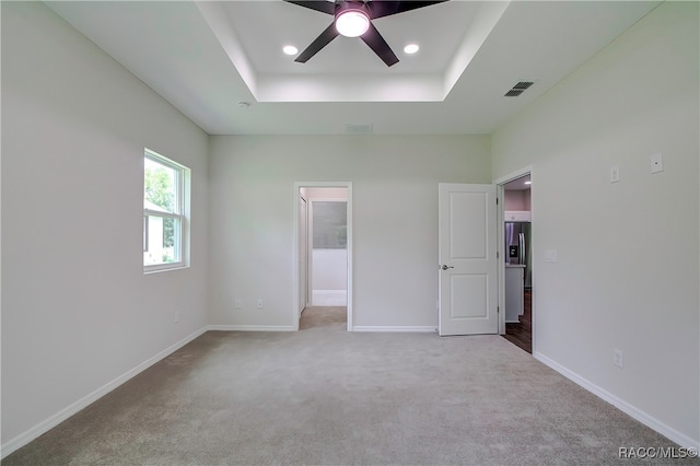 unfurnished bedroom featuring light carpet, ceiling fan, a spacious closet, a tray ceiling, and stainless steel fridge with ice dispenser