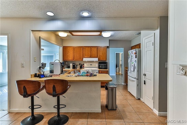 kitchen with brown cabinets, a peninsula, white refrigerator with ice dispenser, light tile patterned flooring, and range