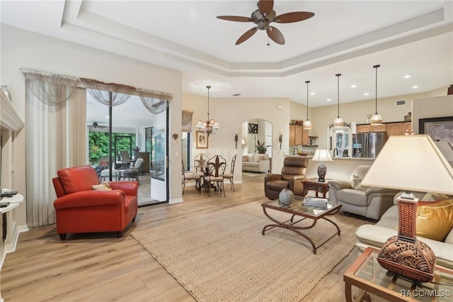 living area with a tray ceiling, ceiling fan with notable chandelier, and light wood finished floors