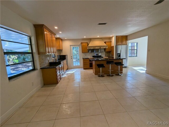 kitchen featuring decorative backsplash, custom range hood, stainless steel appliances, light tile patterned floors, and a kitchen island