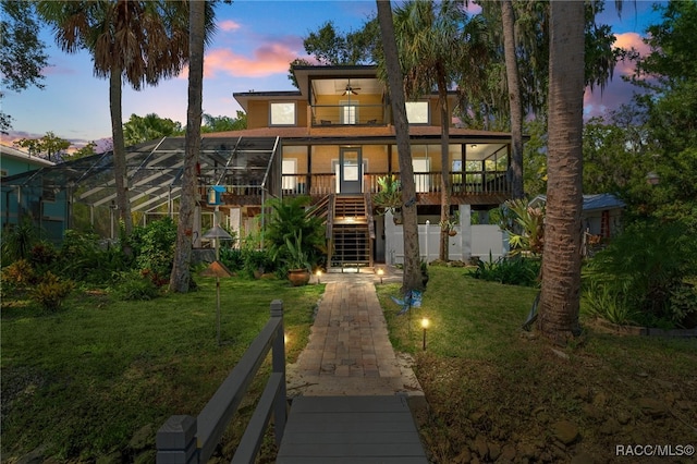 back house at dusk featuring a lawn, glass enclosure, and ceiling fan