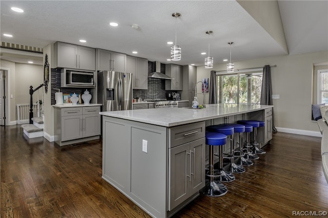 kitchen with wall chimney exhaust hood, dark wood-type flooring, hanging light fixtures, a large island with sink, and appliances with stainless steel finishes