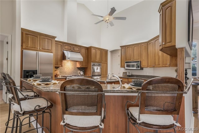 kitchen with tasteful backsplash, ceiling fan, under cabinet range hood, brown cabinets, and stainless steel appliances