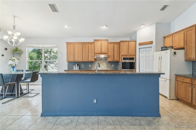 kitchen with stainless steel oven, backsplash, an inviting chandelier, an island with sink, and white fridge