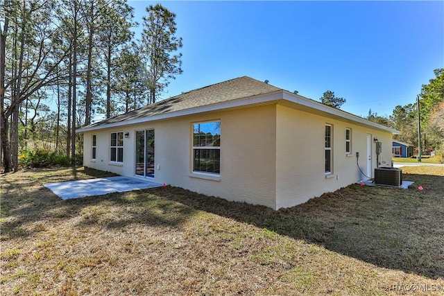 rear view of property featuring stucco siding, a lawn, a patio, a shingled roof, and central AC unit