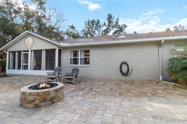 rear view of house with roof with shingles, a patio, stucco siding, an outdoor fire pit, and a sunroom