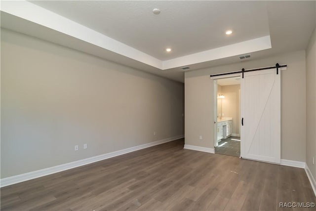 unfurnished bedroom featuring connected bathroom, a barn door, a raised ceiling, and dark hardwood / wood-style floors