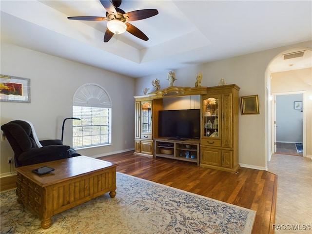 living room with ceiling fan, a raised ceiling, and dark wood-type flooring