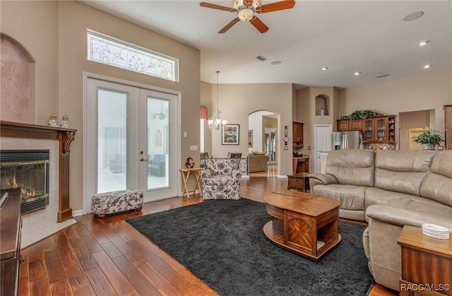 living room featuring ceiling fan with notable chandelier, a high ceiling, a tiled fireplace, dark wood finished floors, and arched walkways