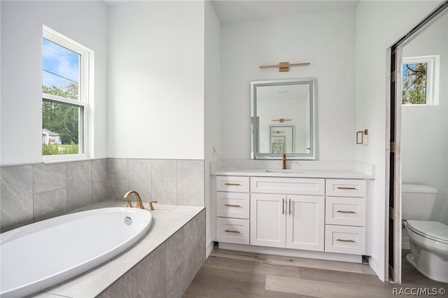 bathroom with wood-type flooring, vanity, a relaxing tiled tub, and toilet