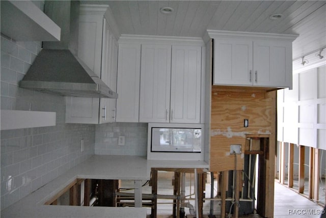 kitchen with white microwave, decorative backsplash, track lighting, wall chimney range hood, and white cabinets