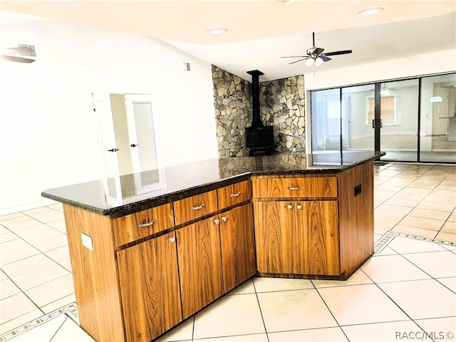 kitchen with a wood stove, dark stone counters, ceiling fan, light tile patterned floors, and kitchen peninsula