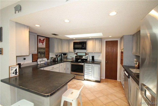 kitchen with dark countertops, a peninsula, gray cabinets, stainless steel appliances, and a kitchen bar