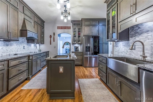 kitchen featuring arched walkways, an island with sink, glass insert cabinets, stainless steel appliances, and a sink