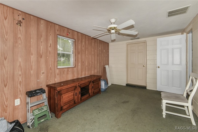 interior space featuring wooden walls, ceiling fan, and dark carpet