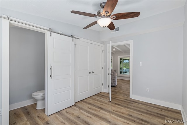 unfurnished bedroom with ensuite bath, ceiling fan, a barn door, light wood-type flooring, and a closet