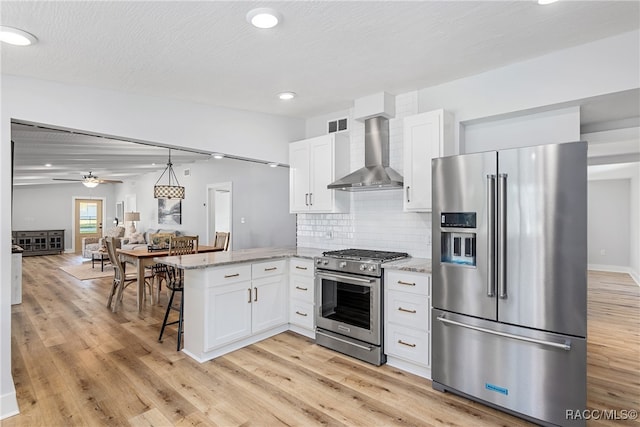 kitchen featuring kitchen peninsula, light stone countertops, stainless steel appliances, wall chimney range hood, and white cabinetry