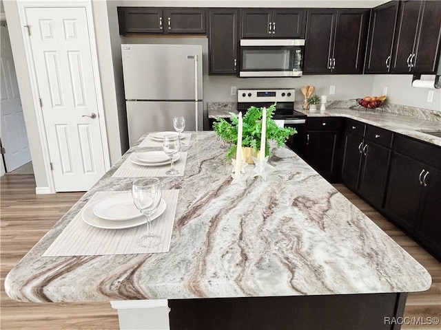 kitchen with dark brown cabinetry, stainless steel appliances, wood finished floors, a center island, and light stone countertops