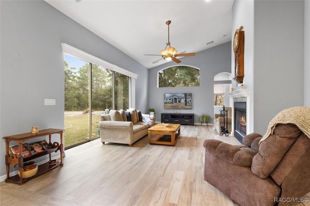 living room with light wood-style flooring, ceiling fan, high vaulted ceiling, a warm lit fireplace, and baseboards