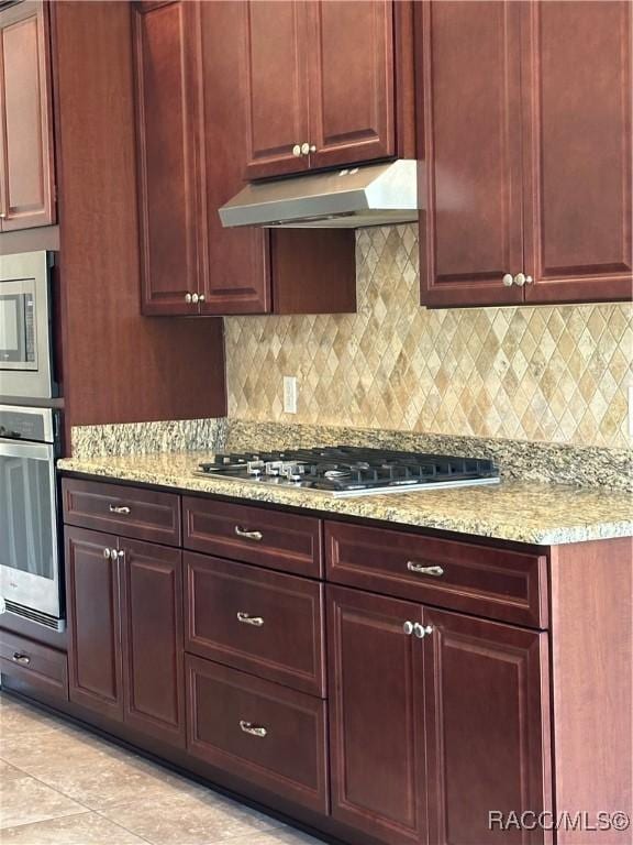 kitchen with reddish brown cabinets, stainless steel appliances, light stone counters, and under cabinet range hood