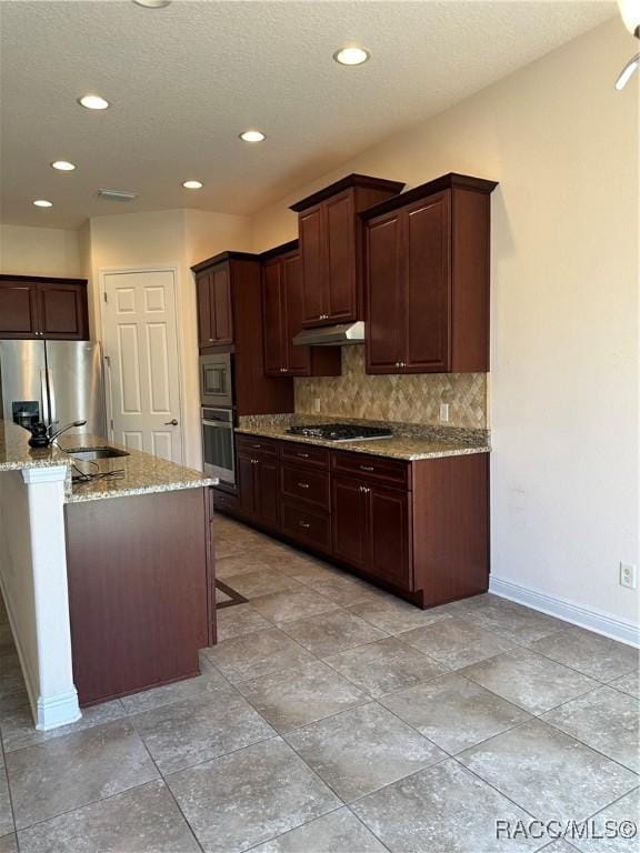 kitchen featuring baseboards, decorative backsplash, appliances with stainless steel finishes, light stone counters, and under cabinet range hood