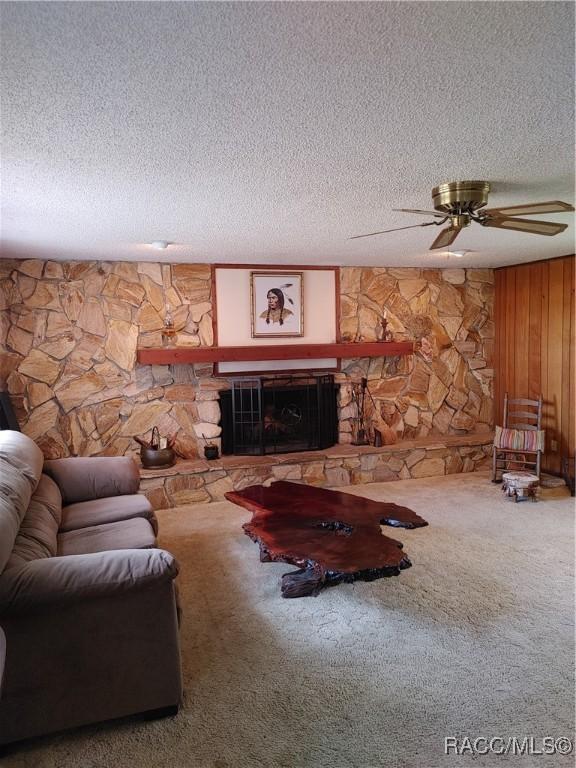 living room with wooden walls, a ceiling fan, a stone fireplace, a textured ceiling, and carpet flooring