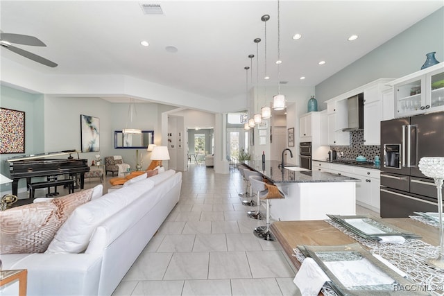 kitchen with appliances with stainless steel finishes, wall chimney exhaust hood, a kitchen island with sink, white cabinetry, and hanging light fixtures