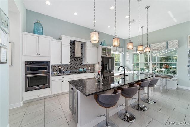 kitchen featuring a kitchen bar, wall chimney exhaust hood, black appliances, a large island with sink, and white cabinetry