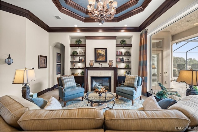 living room featuring built in shelves, a chandelier, ornamental molding, a tray ceiling, and a fireplace