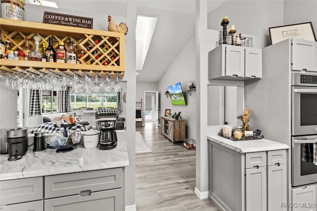 kitchen with light stone countertops, stainless steel double oven, light hardwood / wood-style flooring, vaulted ceiling, and gray cabinets