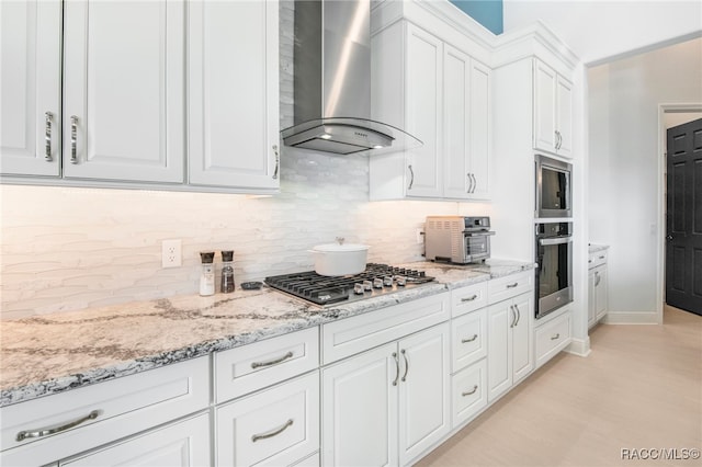 kitchen featuring light stone countertops, white cabinetry, wall chimney exhaust hood, stainless steel appliances, and backsplash