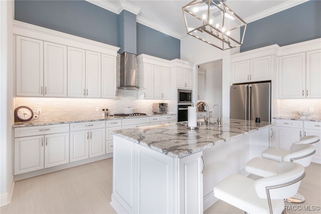 kitchen featuring white cabinets, wall chimney exhaust hood, an island with sink, decorative light fixtures, and stainless steel appliances