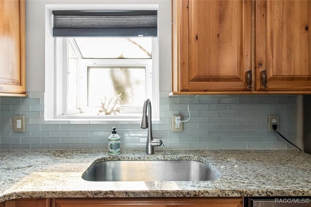 kitchen with brown cabinetry, a sink, backsplash, and light stone countertops