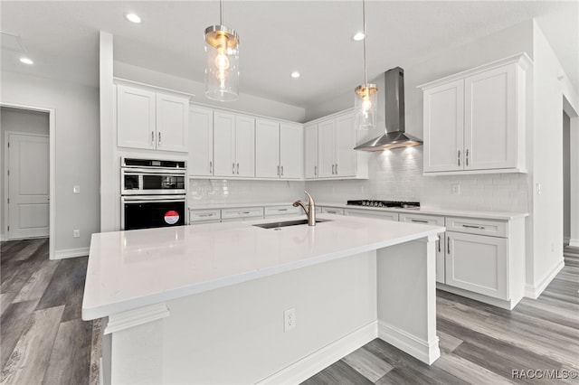 kitchen featuring pendant lighting, white cabinets, a center island with sink, wall chimney range hood, and sink