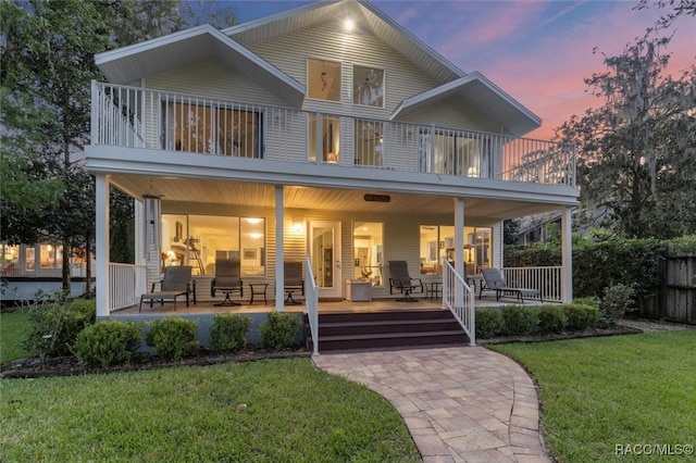 back house at dusk featuring a balcony, covered porch, and a lawn